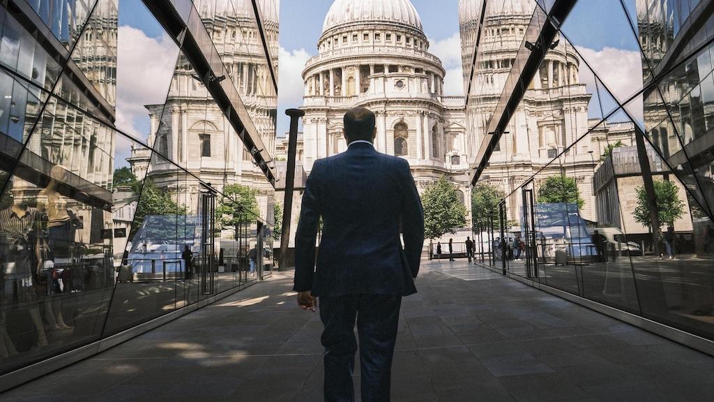 Vishal Marria in a suit walking towards St. Paul's Cathedral, reflected in glass walls on both sides, under a clear blue sky.
