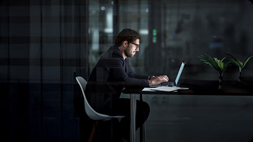 Man working on a laptop at night in a dimly lit office, seated at a desk with a plant and notebook, focused on the screen.