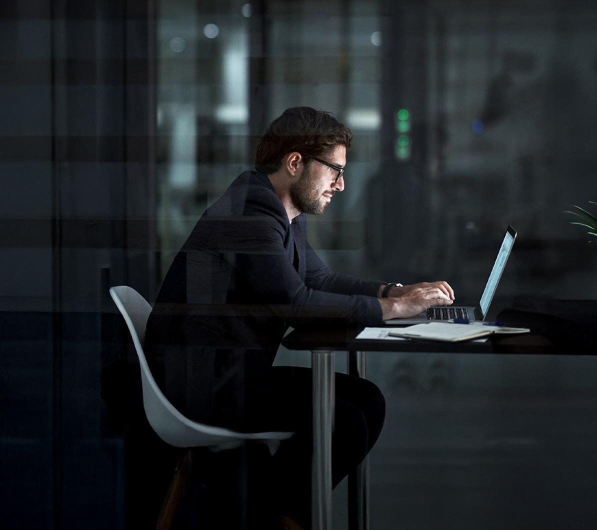 A man in a suit works on a laptop at a desk in a dimly lit office at night, with a notepad and pen beside him.