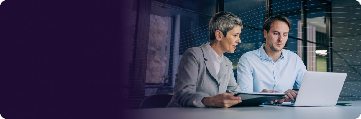 Two people discussing work at a desk with a laptop, one holding a folder. Office setting with a purple gradient on the left side.