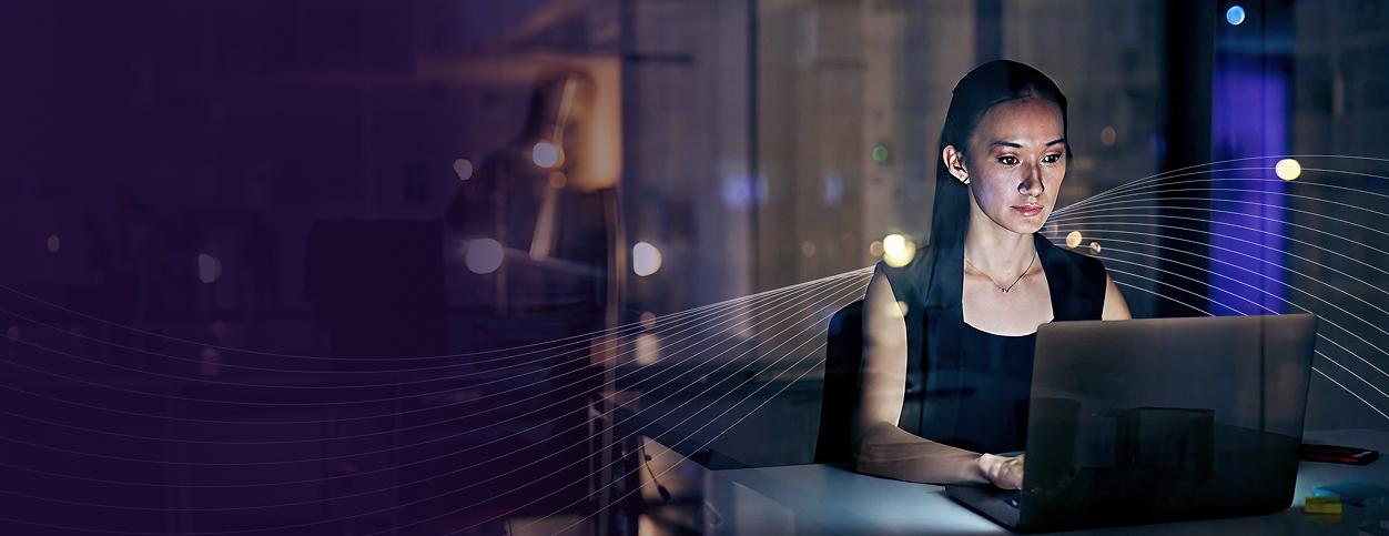 A woman working on a laptop at night in a dimly lit office with abstract lines and blurred lights in the background.