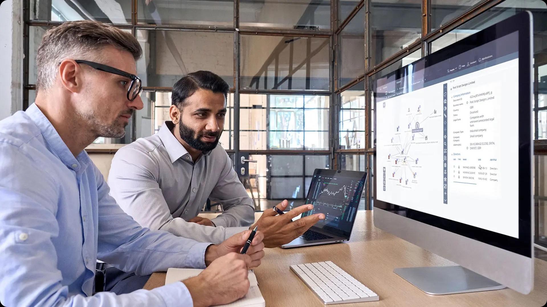 Two men in an office discuss data displayed on a large computer screen. A laptop is open beside them, showing graphs and charts.