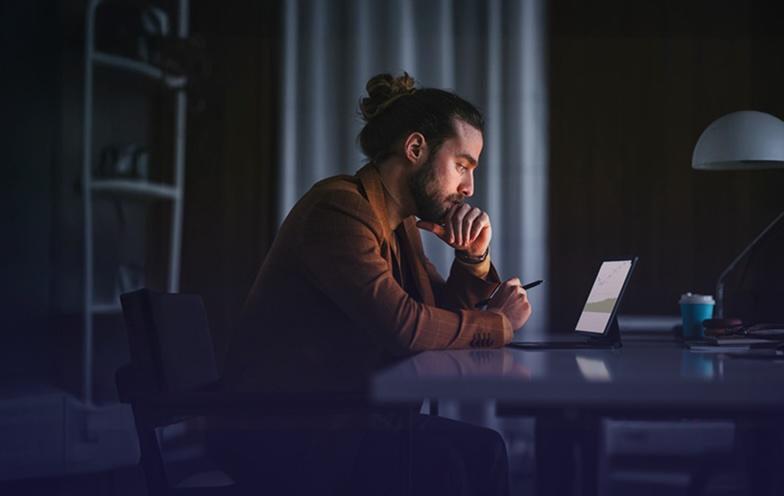 Man with a bun, in a brown jacket, working on a tablet at a desk in a dimly lit room.