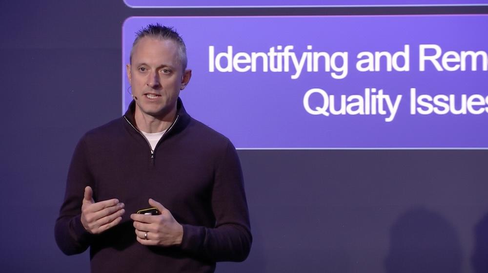 Jamie Hutton speaking on stage about quality issues, standing in front of a presentation slide with a microphone on his cheek.