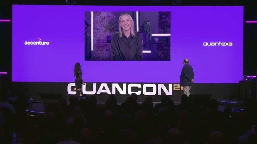 A woman appears on a large screen speaking at a conference, with two people on stage. The screen displays "accenture" and "quantexa" logos.