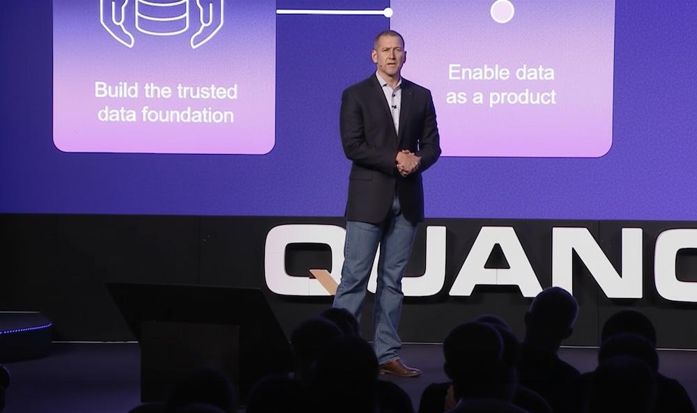 Man in a dark blazer talks on stage, standing in front of a screen displaying tech-related text and diagrams.