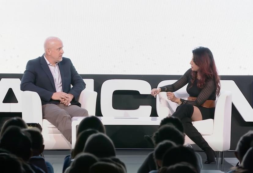 Two people seated on stage with a white backdrop, engaged in conversation. An audience is visible in the foreground.