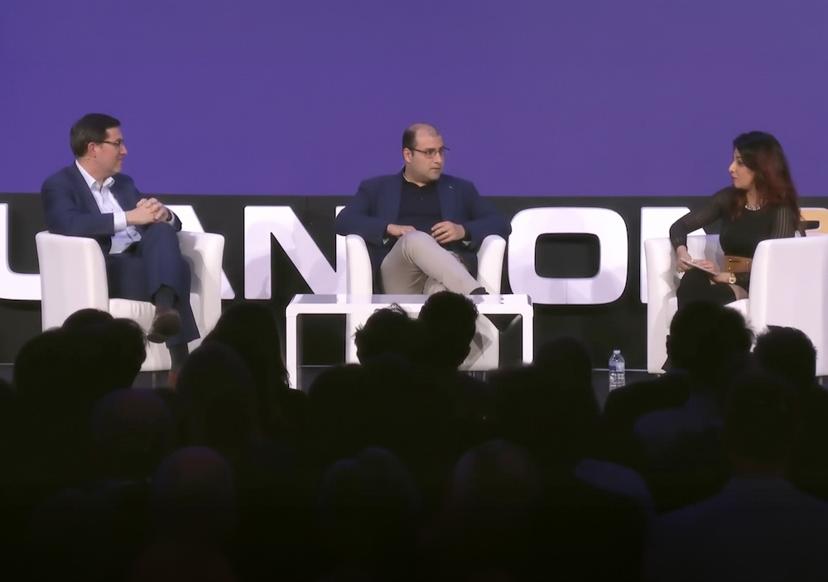 Three people in a panel discussion on stage, seated on white chairs against a purple backdrop, with an audience in the foreground.