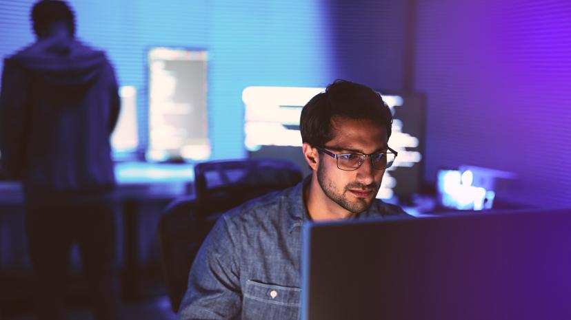A man wearing glasses focuses on a computer screen in a dimly lit room with a purple hue; another person is blurred in the background.