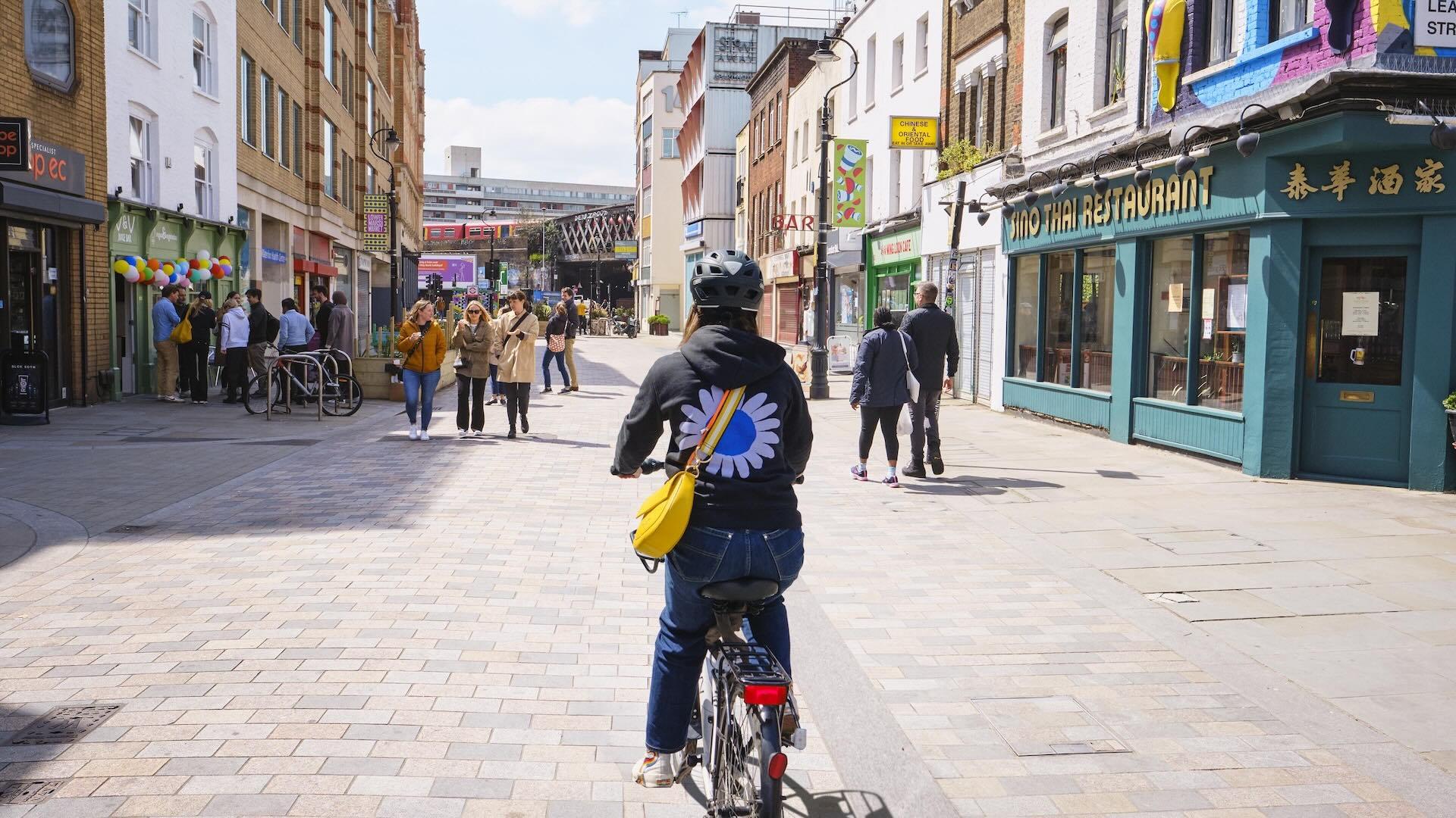 Charlotte is cycling on a pedestrian street lined with shops and restaurants, wearing a black jacket with a daisy design and a yellow bag.
