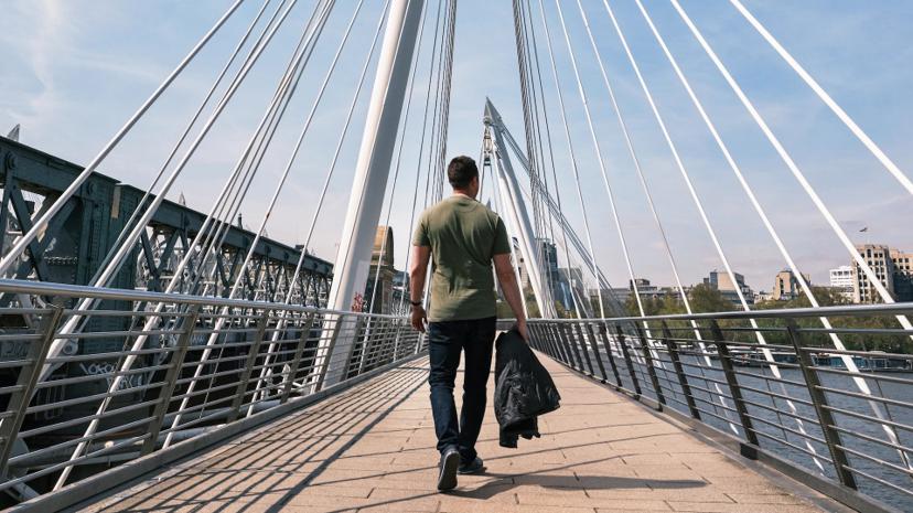 A man walks on a modern suspension bridge, holding a jacket, with city buildings visible in the background under a clear sky.